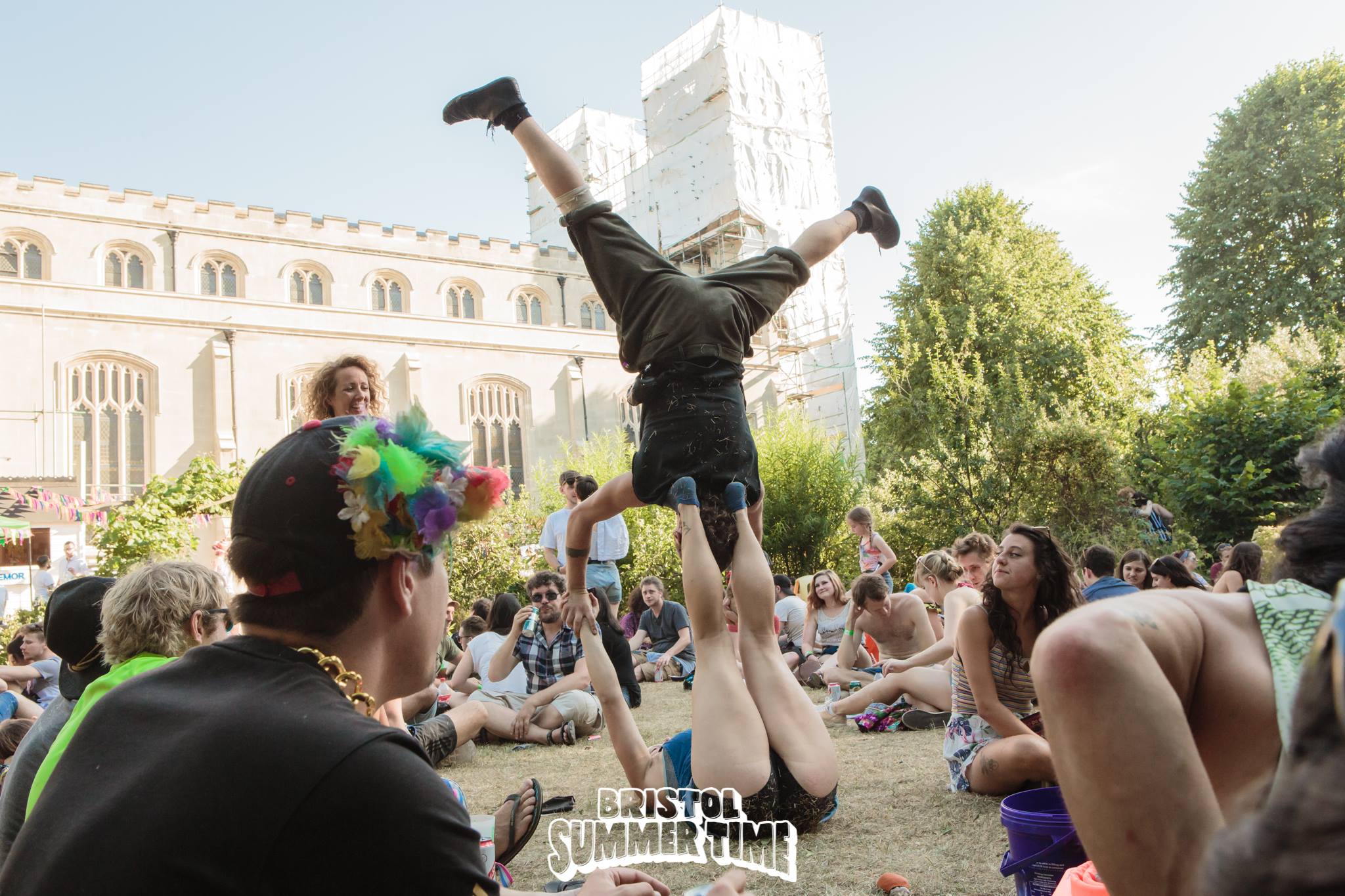 A man upside down balancing on the feet of another person in the garden grounds of Bristol Summer Time festival - watched by a captive audience, with the Trinity Centre, Bristol, in the background