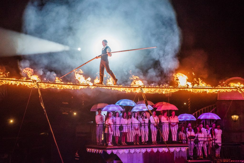 The 2018 Bestival theme was "Circus" - here is a tightrope walker walking over a tightrope that is on fire being watched by the choir dressed in white and holding light-up umbrellas