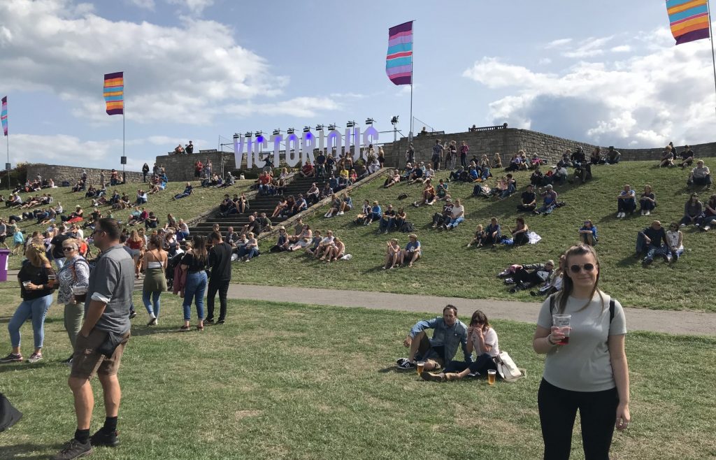 Harriet poses for a photo in the sunshine of the Victorious Case Stage field, with the "Victorious" sign on the hill behind her and festival flags flying in the breeze