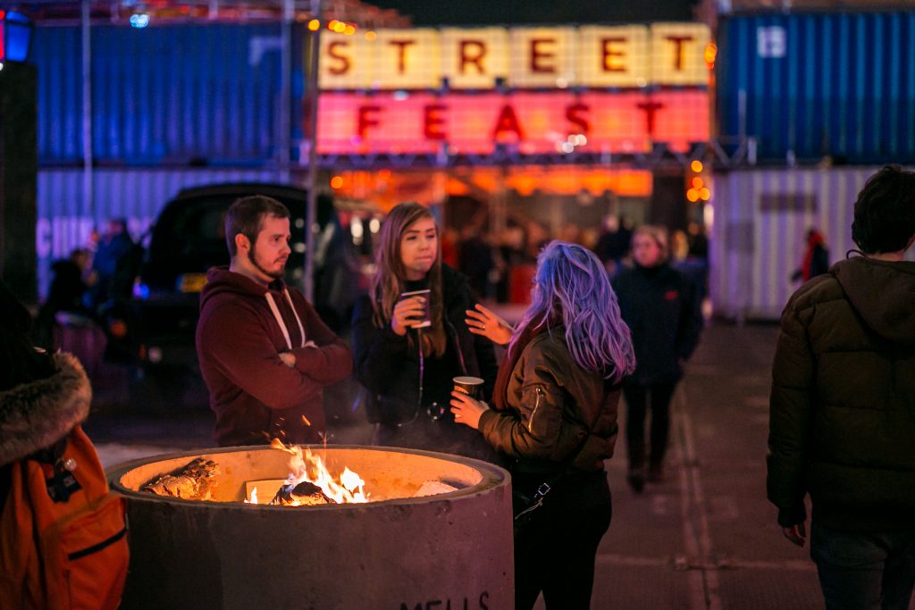 Three friends stood near the front entrance to the Street Feast area which is made from shipping containers. They are holding cups of mulled wine/hot chocolate and are having a conversation.