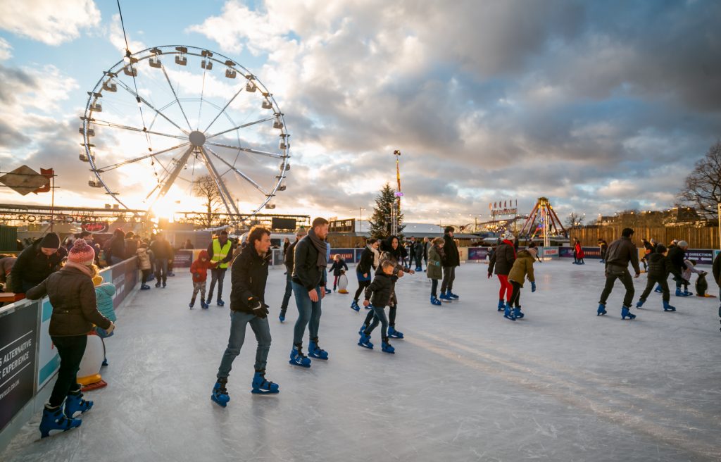 View of the ice rink at Winterville, lots of people ice skating beneath blue but cloudy skies, with the fairground Big Wheel in the background