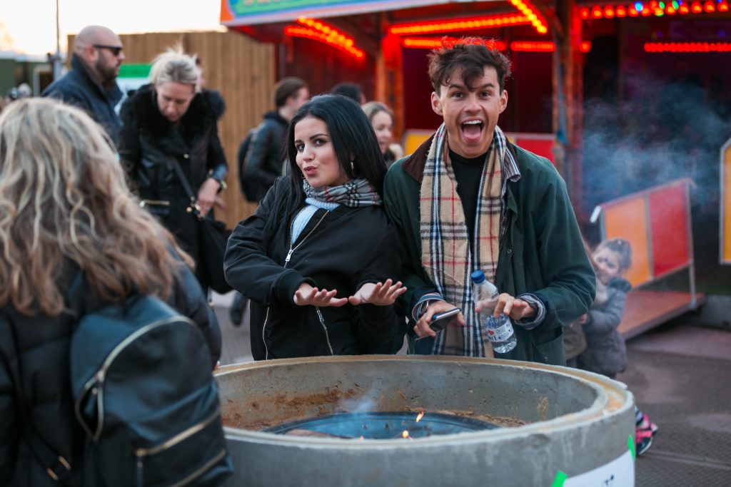 Man and woman stood by a fire pit posing with big smiles at Winterville