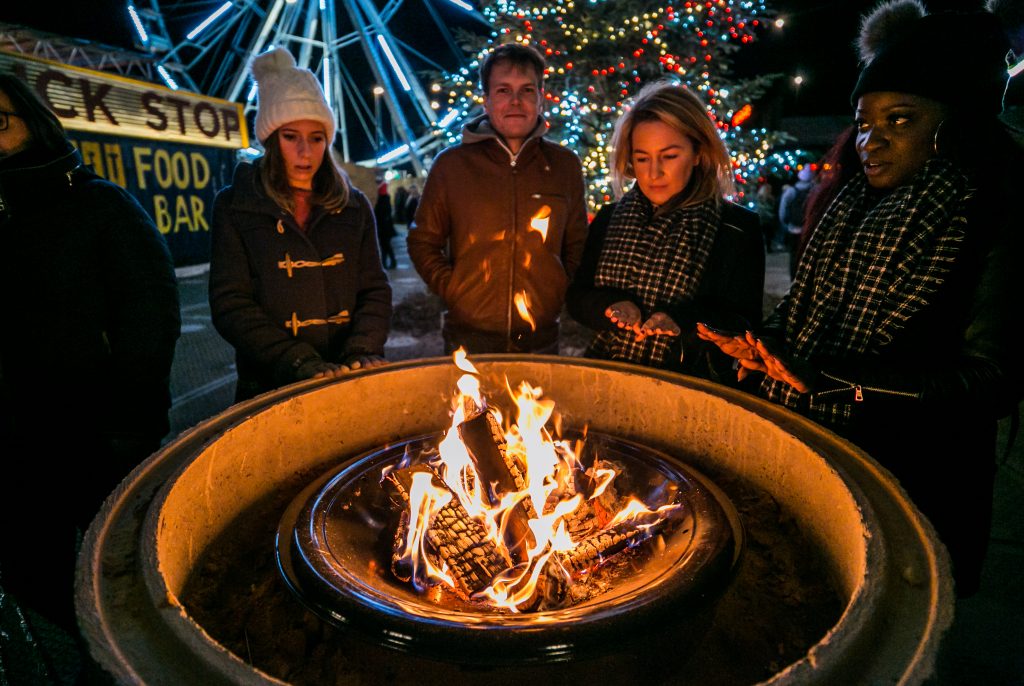 Group of 4 people warming their hands on a fire pit, with the Winterville Christmas tree and big wheel in the background
