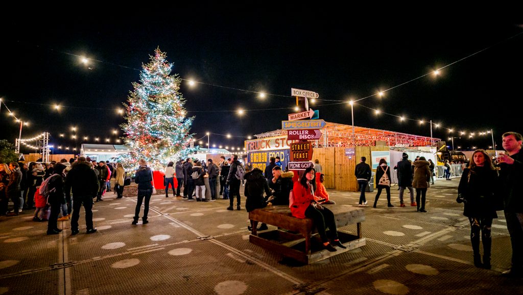 The gorgeous Winterville Christmas tree lit up in all it's glory in the middle of the winter festival site. Lots of people milling around enjoying the atmosphere and christmas lights.