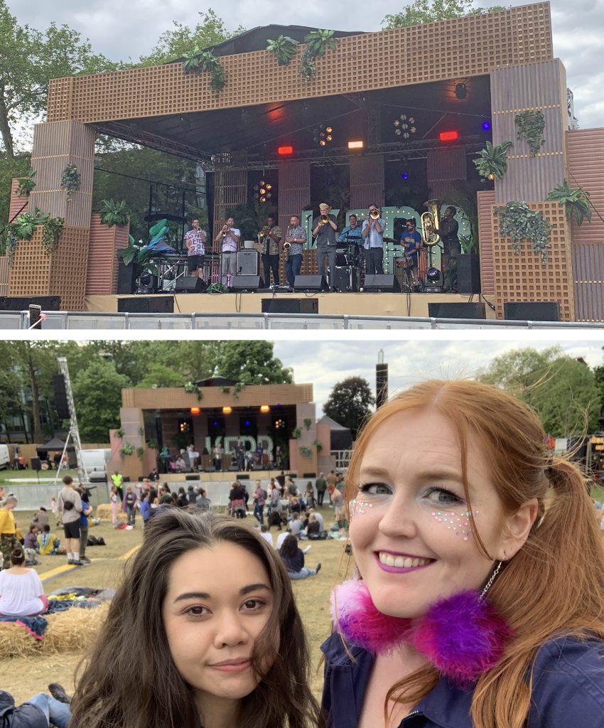 Brasstermind are on the main stage at Jam on Rye Festival, with their many members all player brass instruments. Katey and Jessi smile for a selfie in front of the stage. Jessi has fluffy pink earrings on