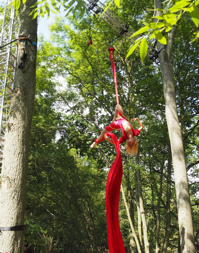 A blonde lady in red is dangling from a (very) high up in the trees frame. She's doing aerial silks and is upside down, holding on with just one leg!