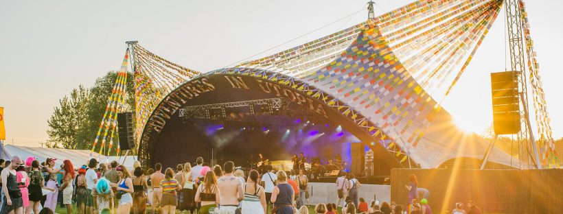 The main stage at Shambala Festival 2019. The stage has an arch other it with "Adventures In Utopia" letters on it and lots of flag bunting. The sun is going down and a band is on stage.