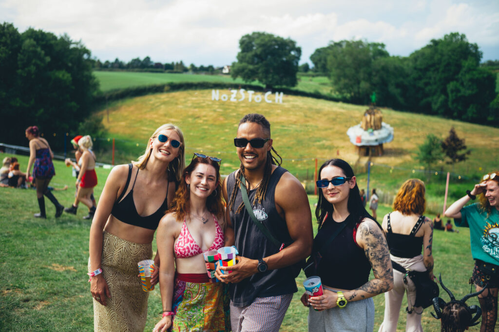 Four friends stand in front of the Nozstock sign high in the field at the festival