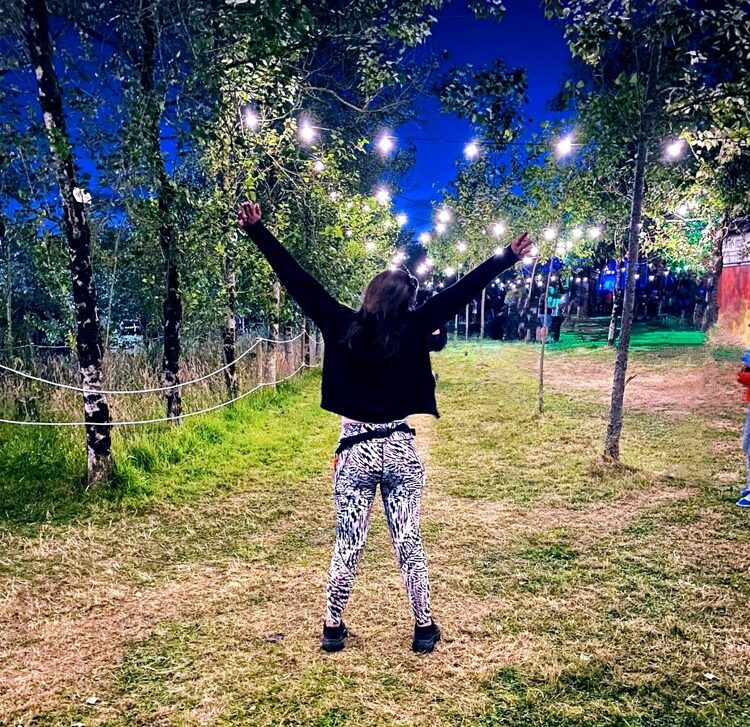 Katt faces away from the camera with her arms in the air - she's wearing funky zebra leggings and is standing between some trees with twinkly lights. She's clearly excited for the night ahead at Leo!