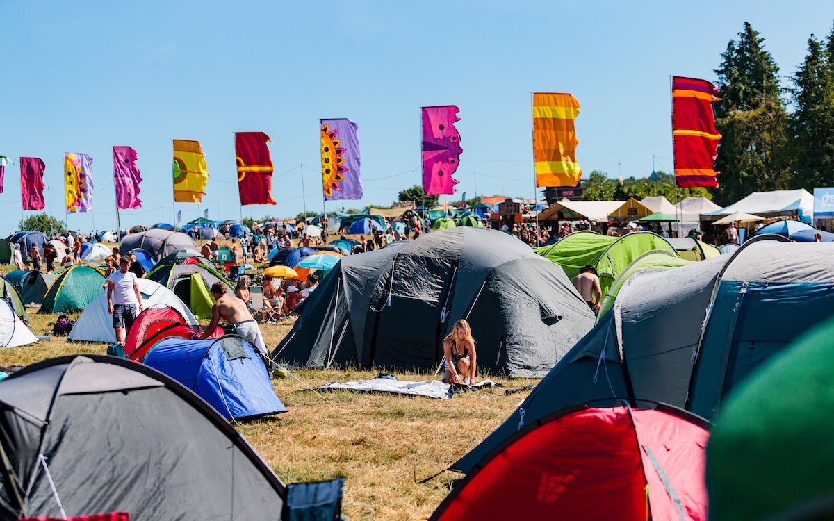 A sunny day at Boomtown Fair in the Camp Orchid campsite. There are tents in the foreground with lots of space between them, a blue cloudless sky, and multicoloured flags blowing in the wind.