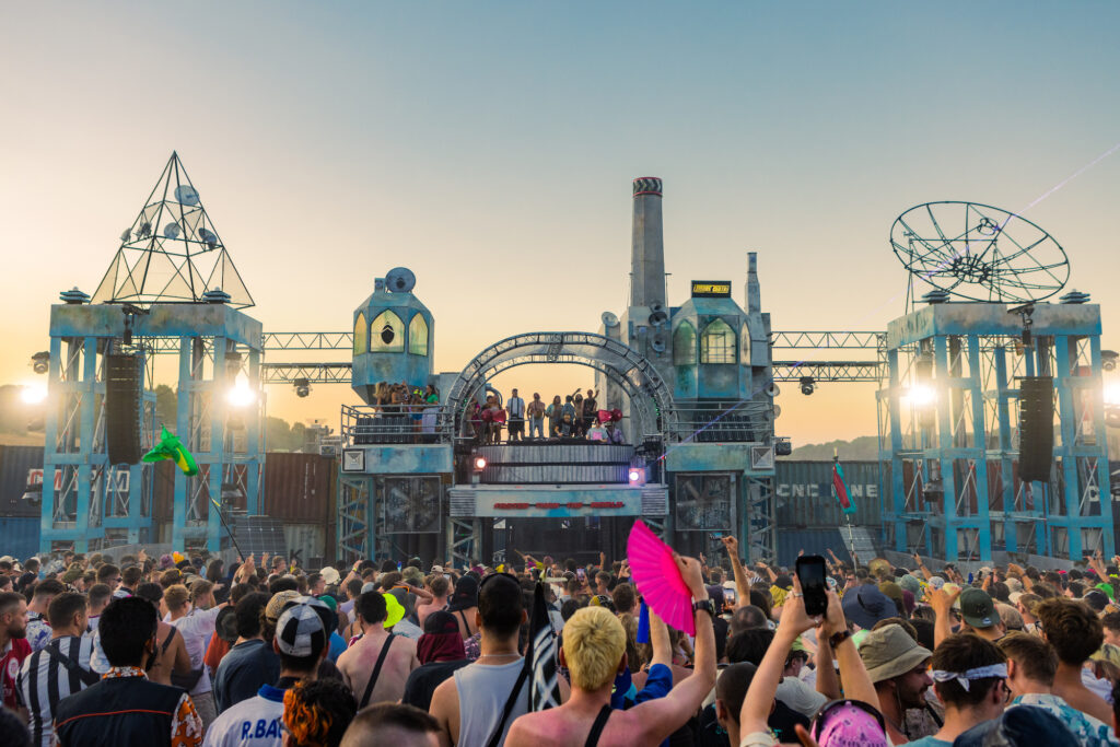 The Grid stage in Area 404 at Boomtown Fair - a large metal structure with a pyramid structure on one side and a dish-shape structure on the other. It looks like a mad signal station for gamma rays (or something). The sun is setting, and the crowd are all facing the stage which has lots of dancers on the top.