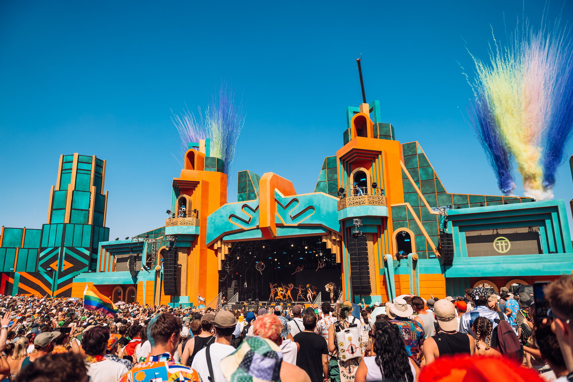 A bright gorgeous image of the brand new Grand Central stage at the Boomtown Fair 2022 Chapter One. It's a huge stage painted bright orange and teal blue, incorporating the Boomtown logo. There are smoke cannons going off behind the stage like daytime fireworks, in front of a bright blue cloudless sky.