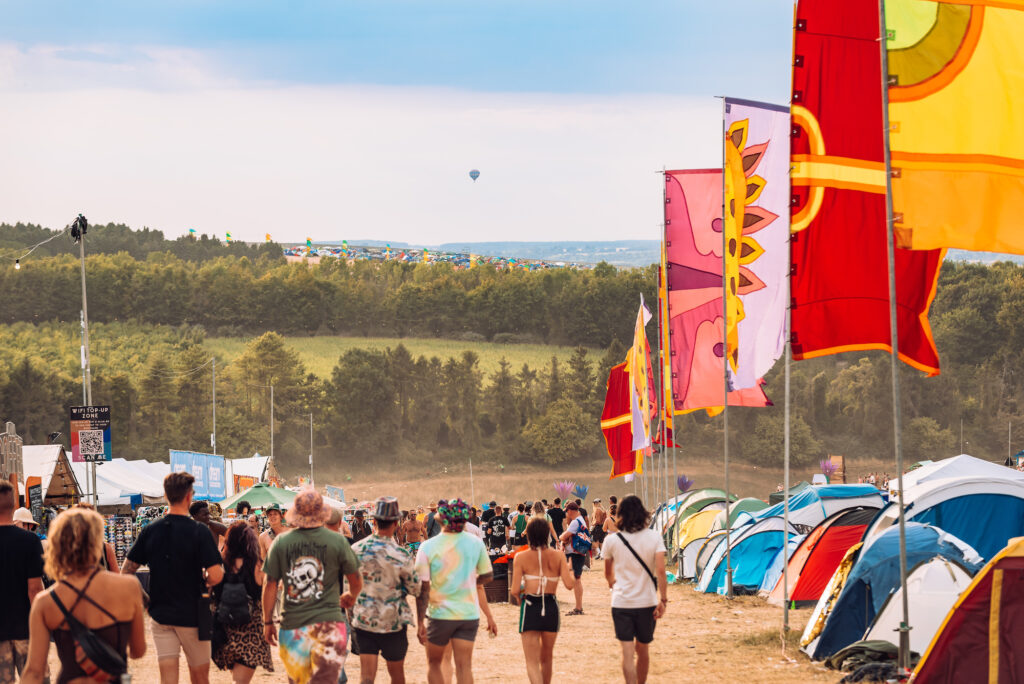 The view down the Hippie Highway at Boomtown Fair Chapter One. Tents are on the right hand side of the picture, which faces down the main path. Festival goers are all walking down the hill towards the main festival area. There are shops on the left hand side, and in the distance on the horizon is a hot air balloon.