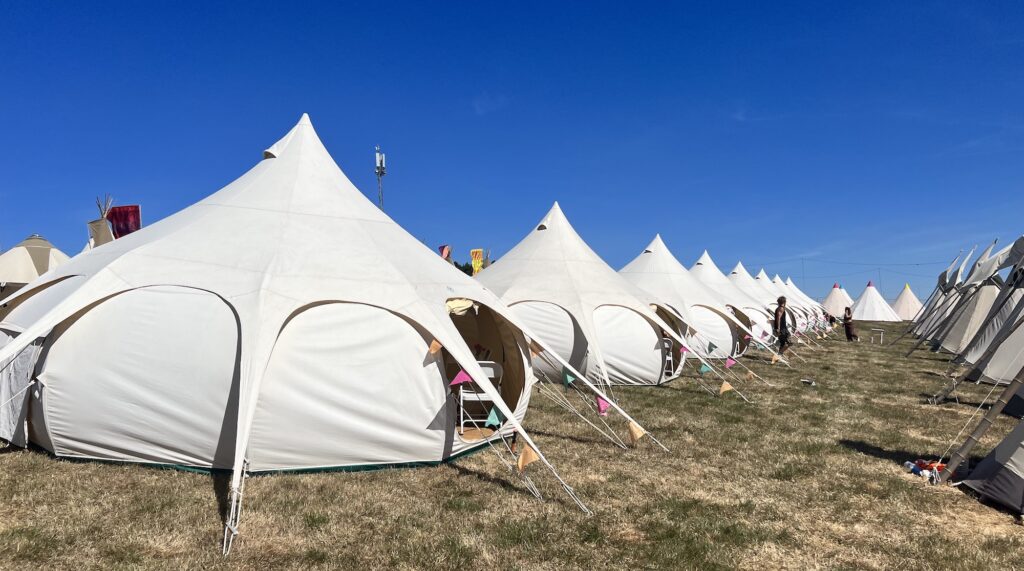 A beautiful sight - a row of 8 large white canvas Lotus Belle tents on grass, each with coloured bunting by the door, with a rich blue sky above. This is the luxurious paradise that is the Portobello Tents area in Camp Skylark at Boomtown Fair.
