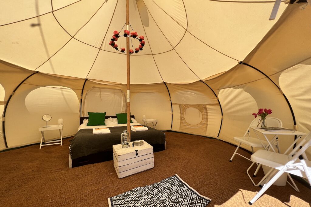 A peek inside one of the large Lotus Belle tents, from Portobello Tents. These tents are curved outwards and very tall, allowing you to walk in and stand up the whole way round the tent. There is so much space! You can also see the double bed with green cushions, a fluffy white towel, and a lovely chandelier with fake roses on it. There's also a rug floor, a table and chairs, and bedside tables with lamps on them.