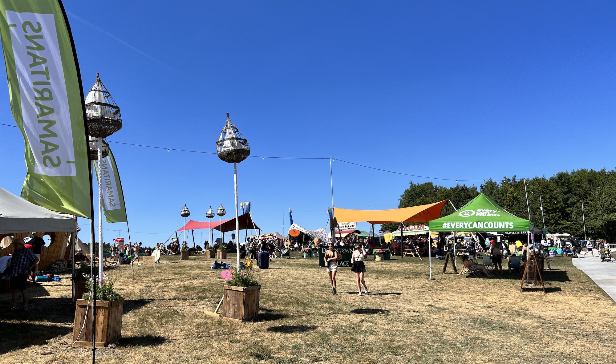 The Downtown Campsite Village Hub area at Boomtown Fair 2022. There are shaded areas under colourful tarps, festival goers strolling around, food trucks in the background. There is lots of space, and the ground is almost completely free of rubbish. You can see the beercan recycling point to the right of the image.