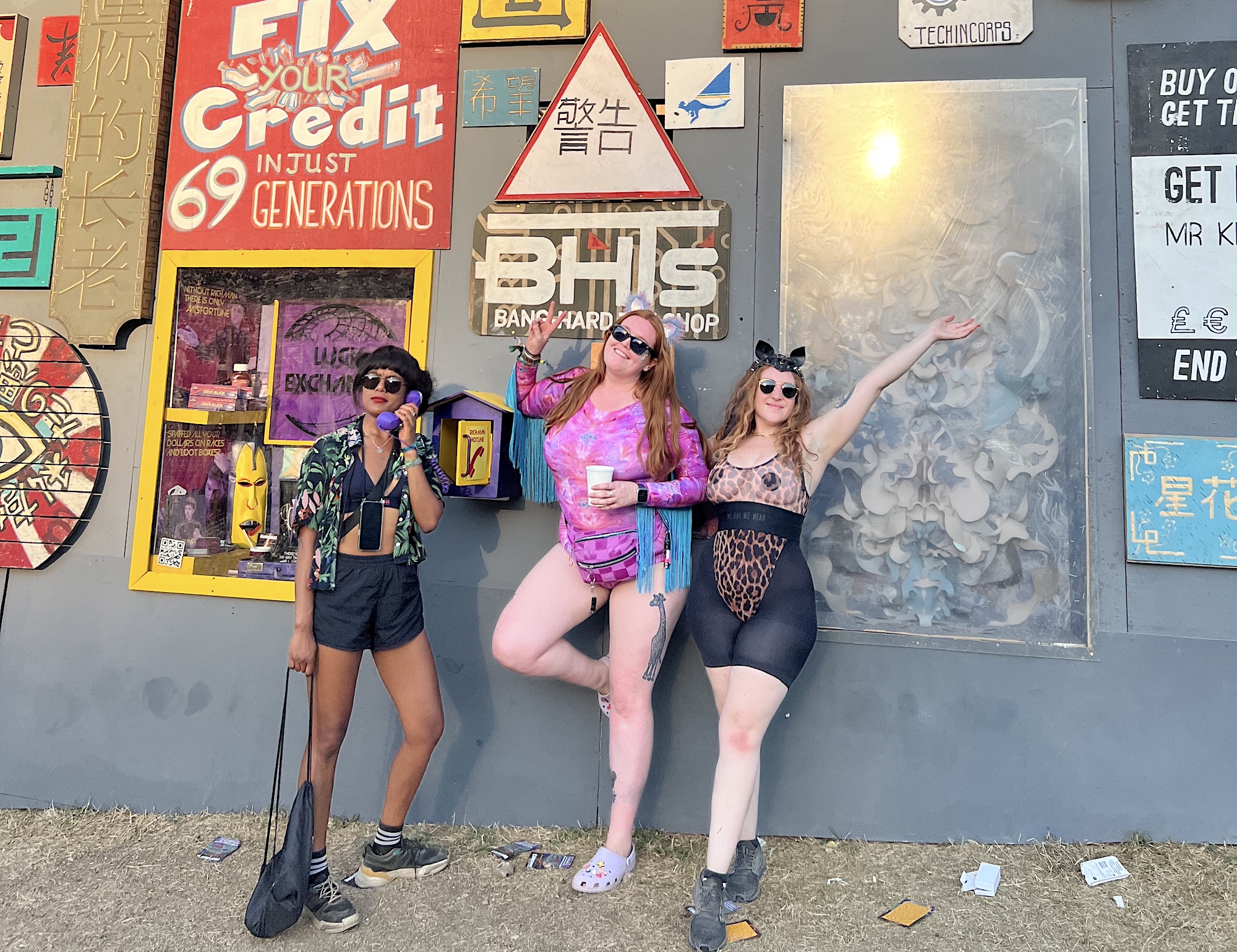 Alex, Laurie and Jessi are stood in front of a wall in Metropolis, which is covered in posters for various 'services' in Boomtown. Laurie is answering a landline phone that is on the wall (like a payphone). Jessi and Alex are both in festival patterned bodysuits, and if I do say so myself, us girls look pretty fly!