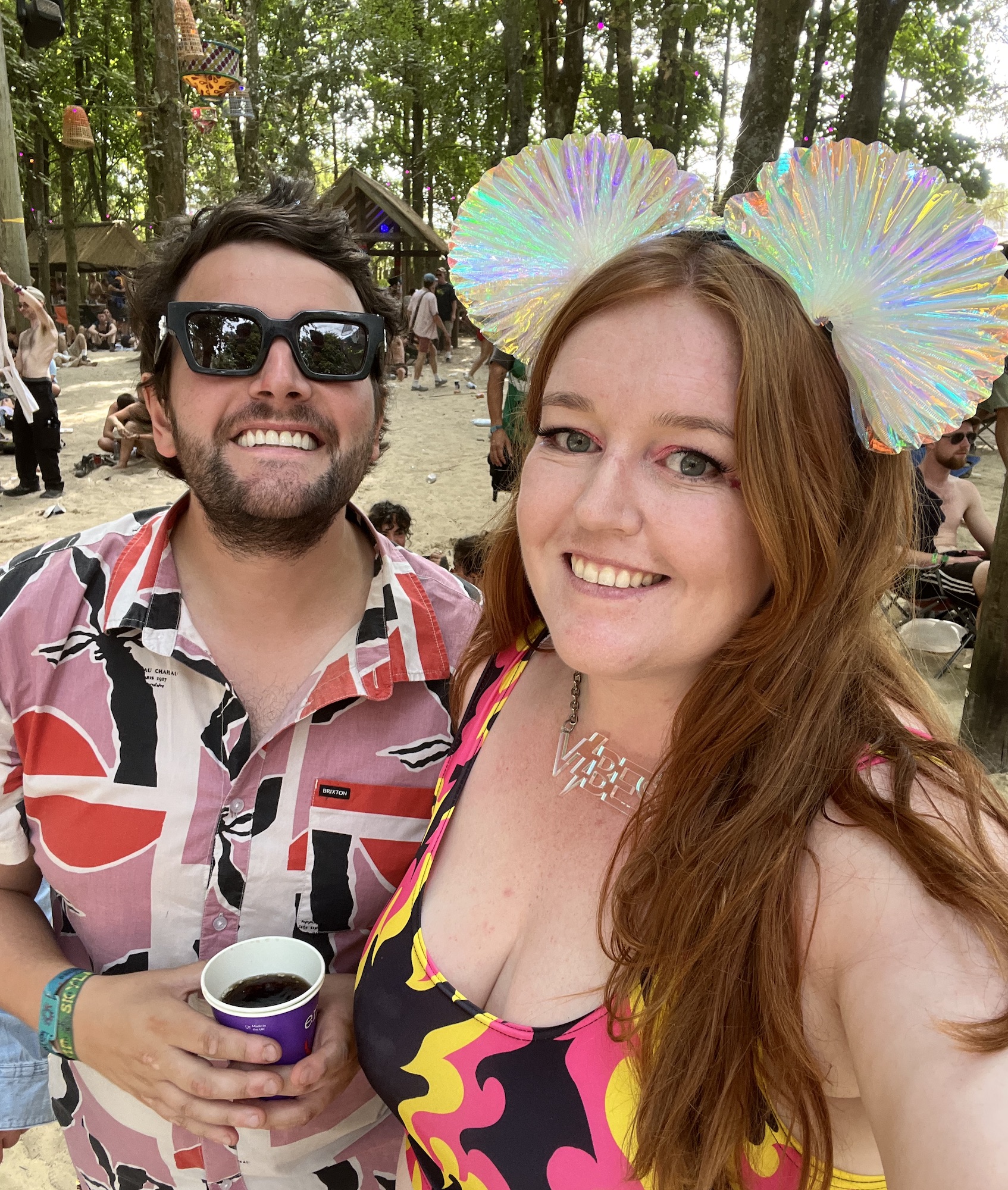 A selfie of Jessi and Ben, taken in the Hidden Woods at Boomtown Fair. It's lovely a shady. Ben is wearing a pink and red graphic shirt and chunky sunglasses, and Jessi has an iridescent headpiece on, an a pink flame print crop top.