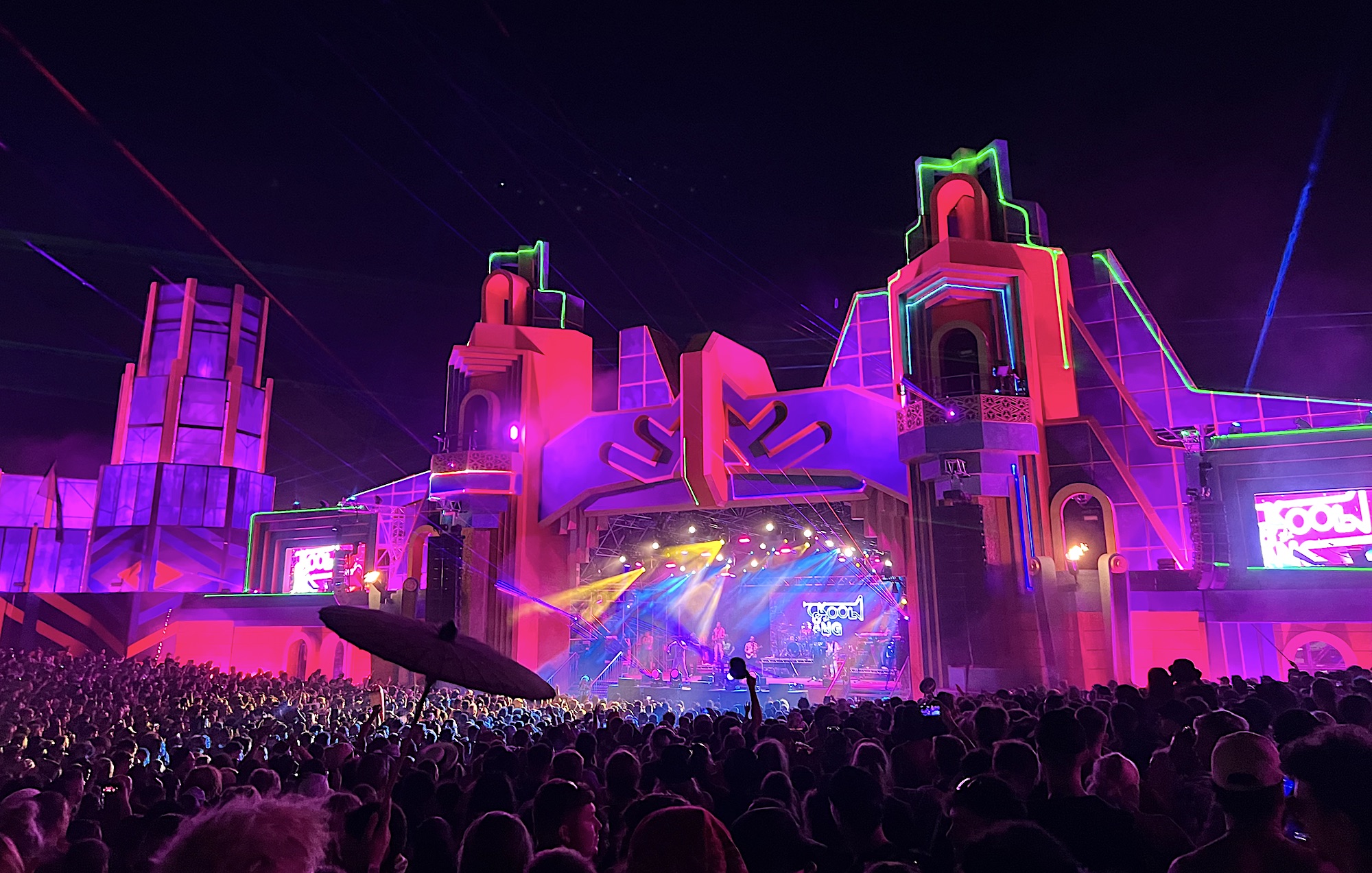 The Grand Central Stage at Boomtown Fair Chapter One, at night, glowing a pinkish red and purple, with the projection mapped lasers illuminating sections of the stage. Kool and the Gang are on stage and the crowd is lit up from the stage lights.