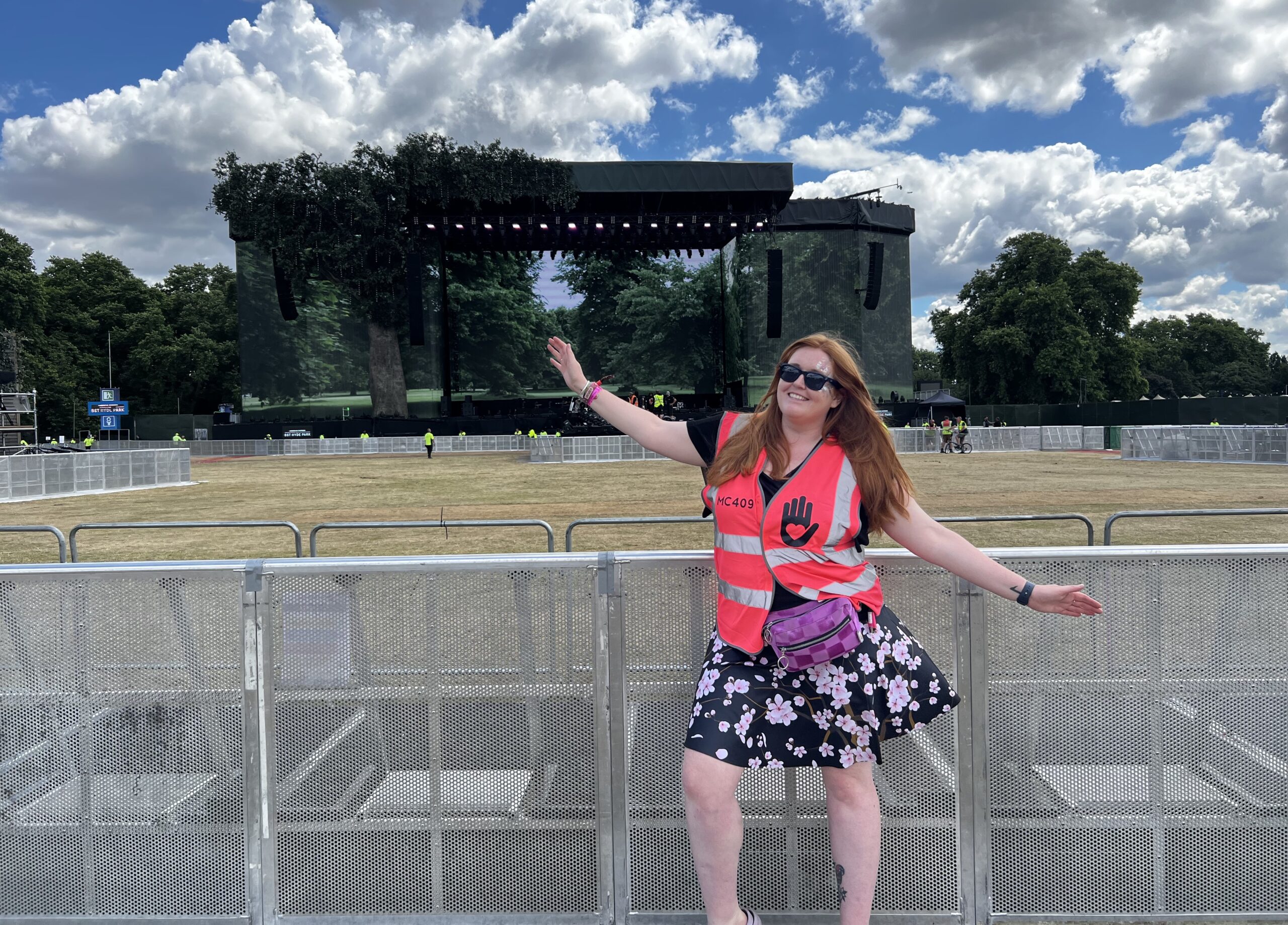 Jessi is stood in front of the huge BST Hyde Park main stage before the gates opened, so the area is empty except for some security. She is wearing a floral dress with her My Cause UK volunteer tabard over the top, and her pink bumbag.