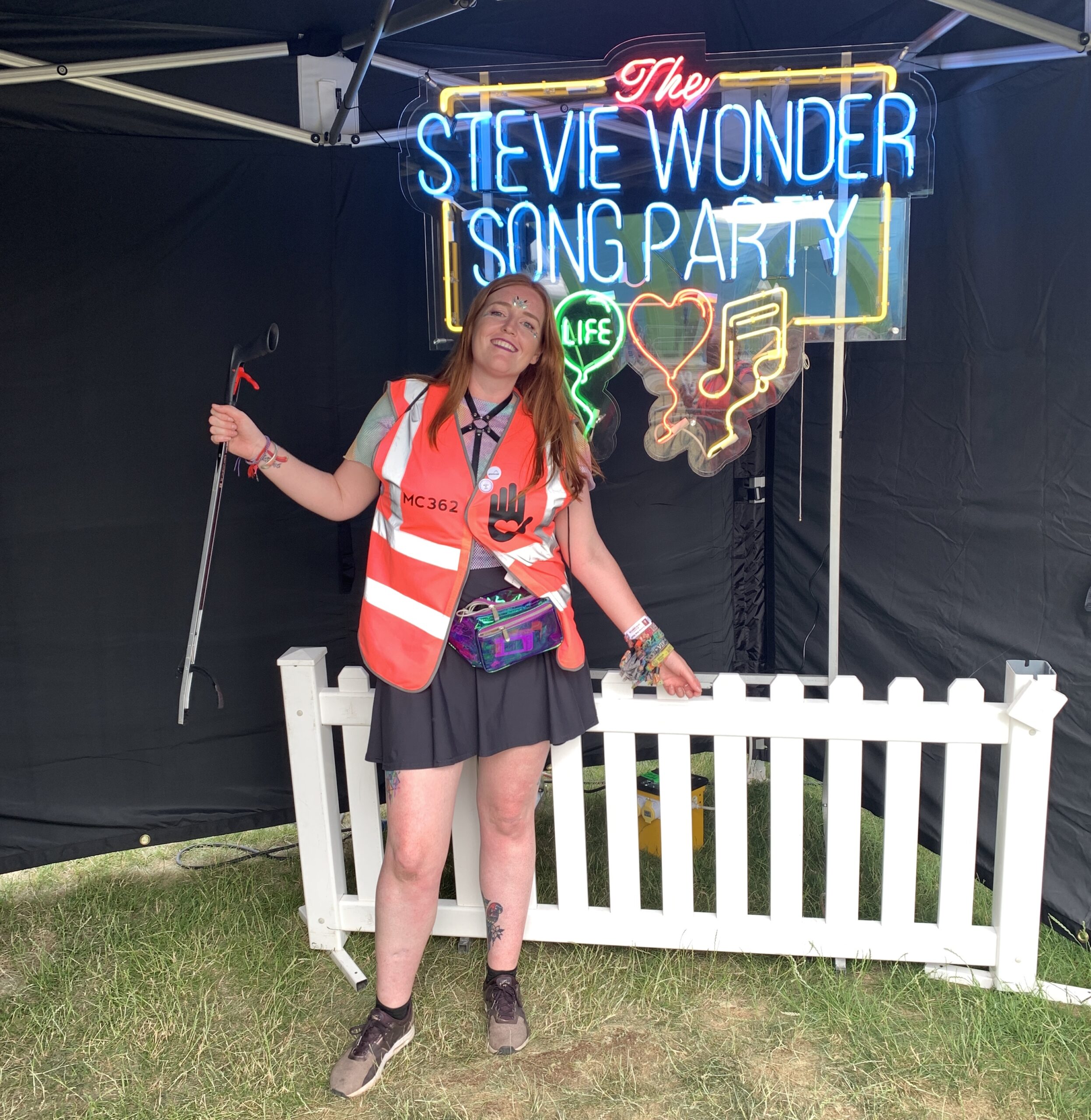Jessi stands in front of a neon sign that says 'Stevie Wonder's song party'. She's ready to go volunteering on the recycling crew and s holding a litter picker.