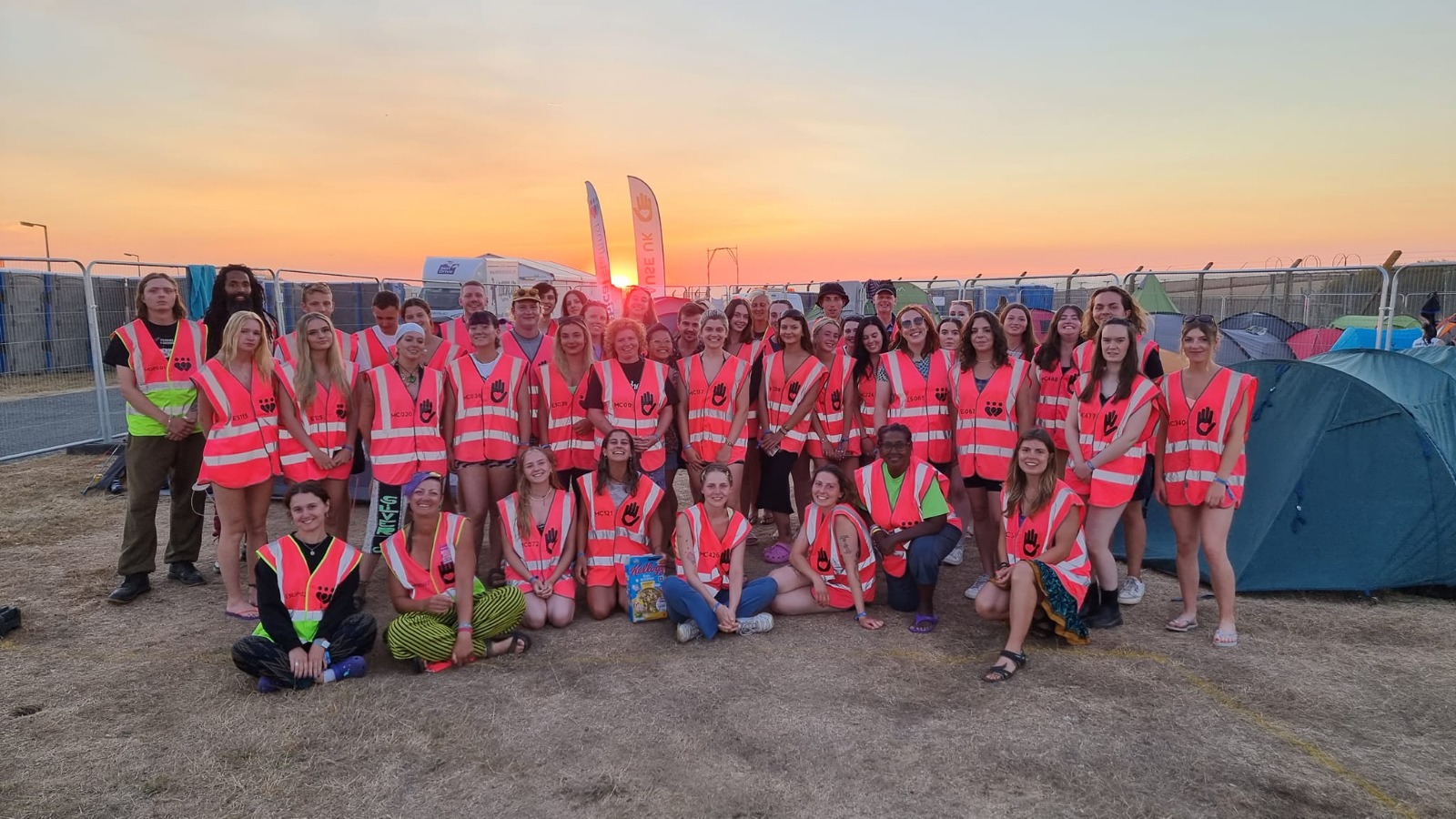 A group of 30 plus smiling volunteers all wearing their bright pink high vis tabards with tents in the background, a My Cause UK flag, and a beautiful sunset behind them.