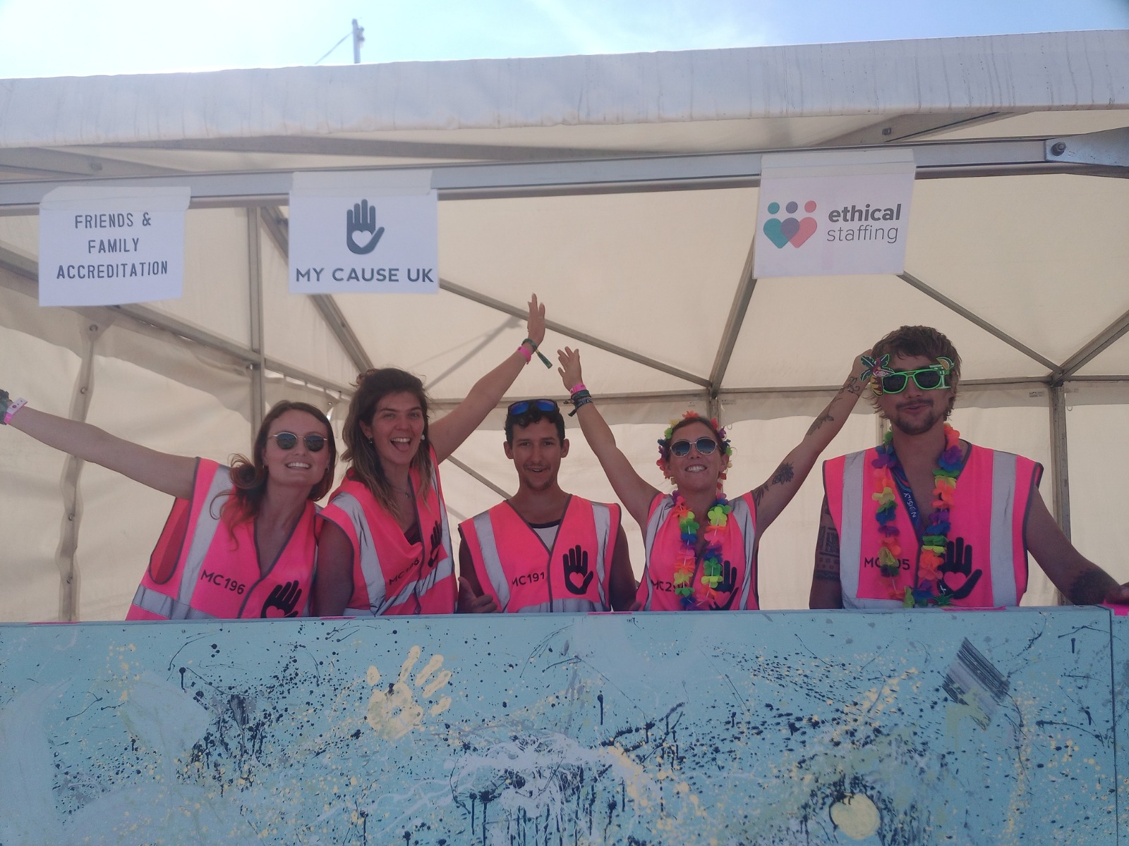5 volunteers are stood in a box office waiting to give accreditation. Two have their arms raised in excited welcome, and two are wearing floral garland necklaces over their neon pink volunteer tabards.