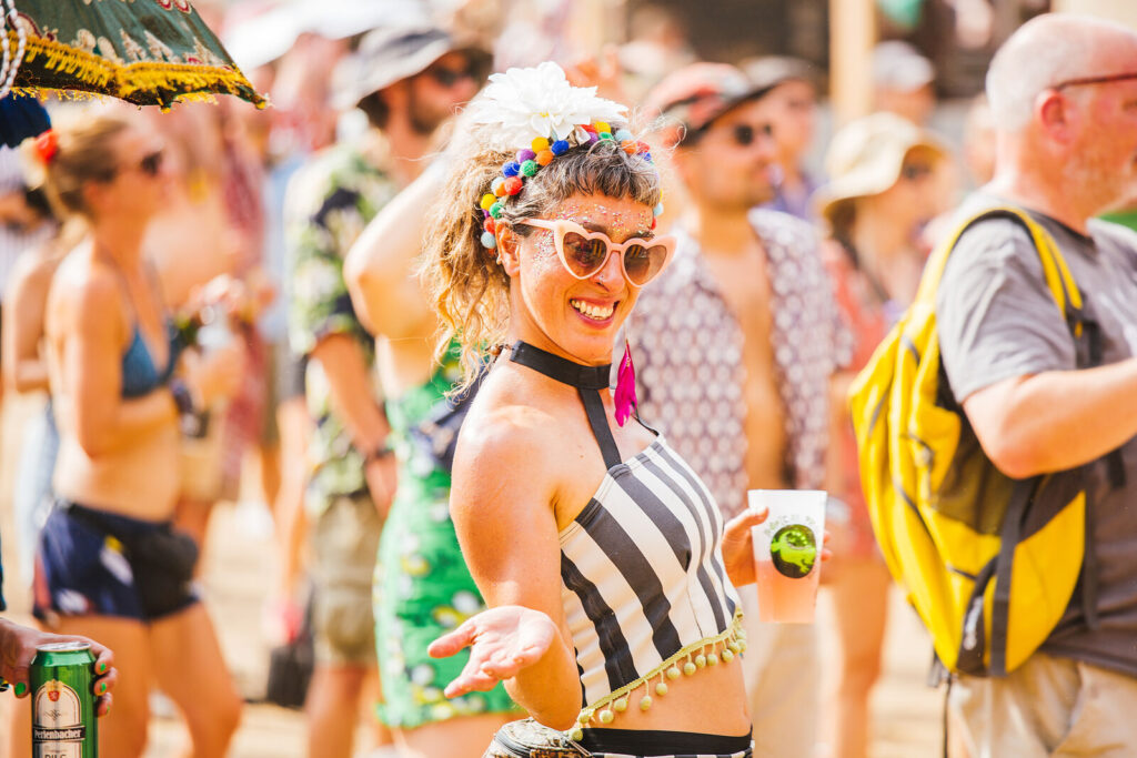 A gorgeous reveller at Boomtown Fair Chapter One: The Gathering. She is smiling at the camera, with pink heart shaped sunglasses, pom poms in her hair, a striped tube top on, and a pint glass in her hand.