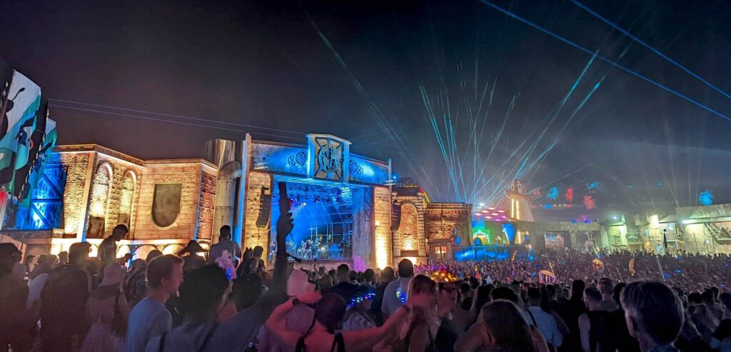 A nighttime view of the Engine House stage in Copper County at Boomtown Fair 2022. There is a huge crowd, and blue lasers are blasting into the sky. There's lots of colours and amazing lights.
